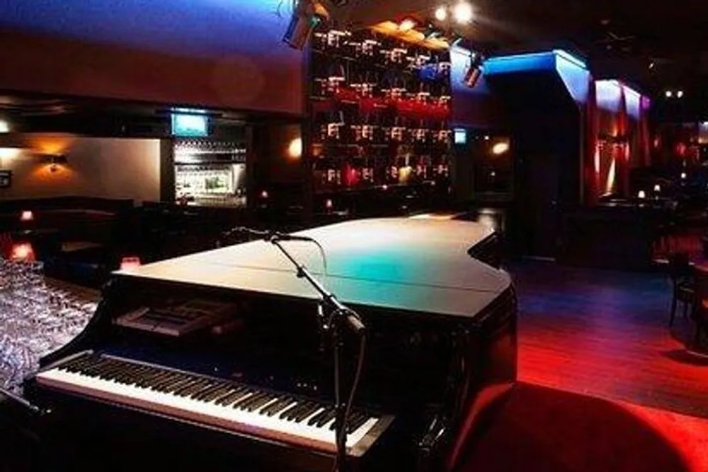 The image shows a dimly lit jazz club with a grand piano in the foreground and a bar with colorful shelves in the background