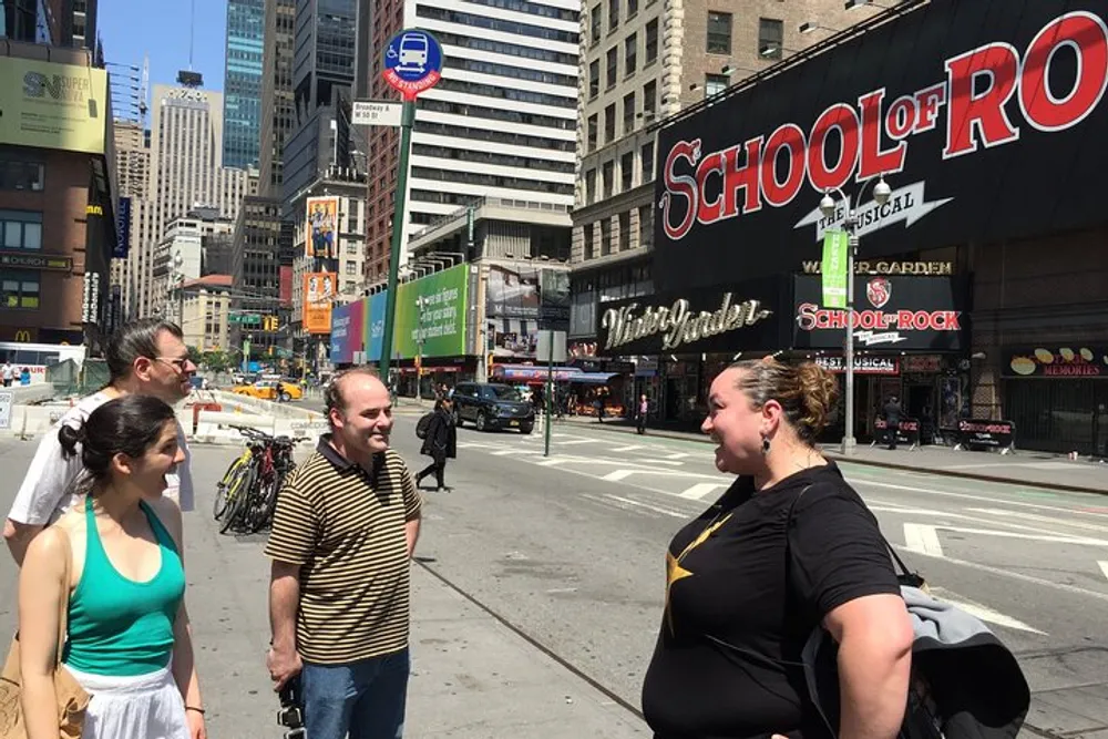 A group of people are chatting on a sunny street corner with a backdrop of the School of Rock Broadway musical billboard and urban scenery