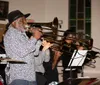 A man in a hat plays the trombone with a band of brass musicians in the background