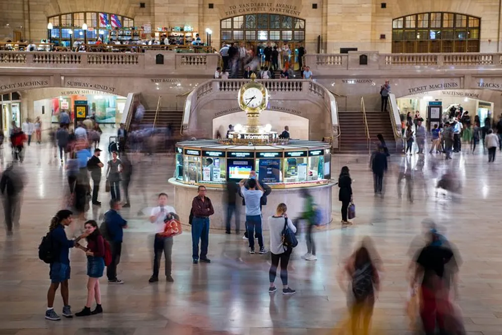 The image captures a bustling interior of a grand train station with motion-blurred figures of commuters and a prominent information kiosk with a clock on top indicating a scene of dynamic transit
