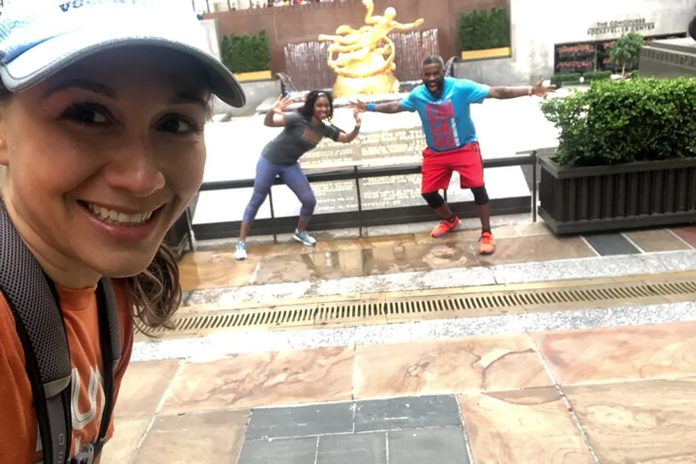 A person is taking a selfie while two others pose playfully in front of a decorative fountain all three wearing athletic attire and smiling