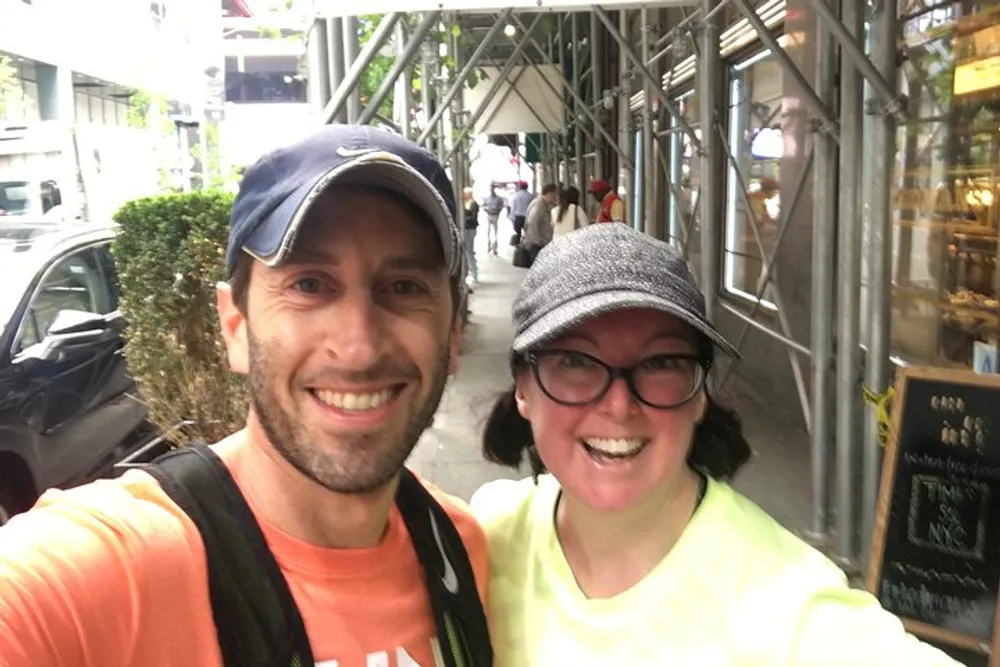 Two people are smiling for a selfie on a city sidewalk where scaffolding is visible in the background