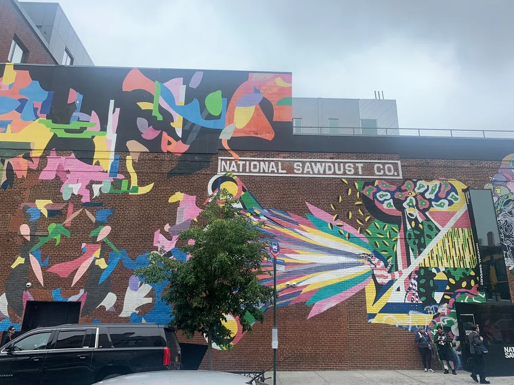 The image showcases a vibrant abstract mural on the exterior of a brick building with a sign that reads National Sawdust Co and several people are present near the entrance
