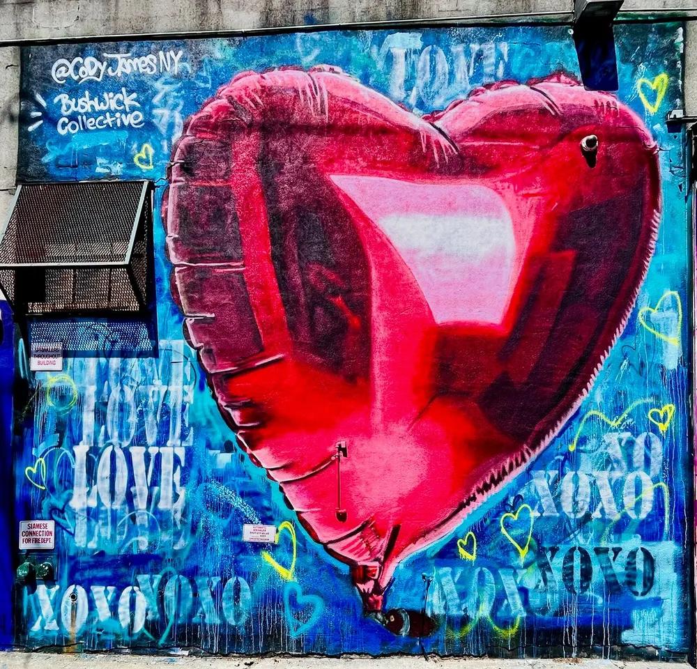 The image shows a vibrant street art mural featuring a large red heart balloon with a patch set against a blue graffiti wall with multiple love-related inscriptions and hearts