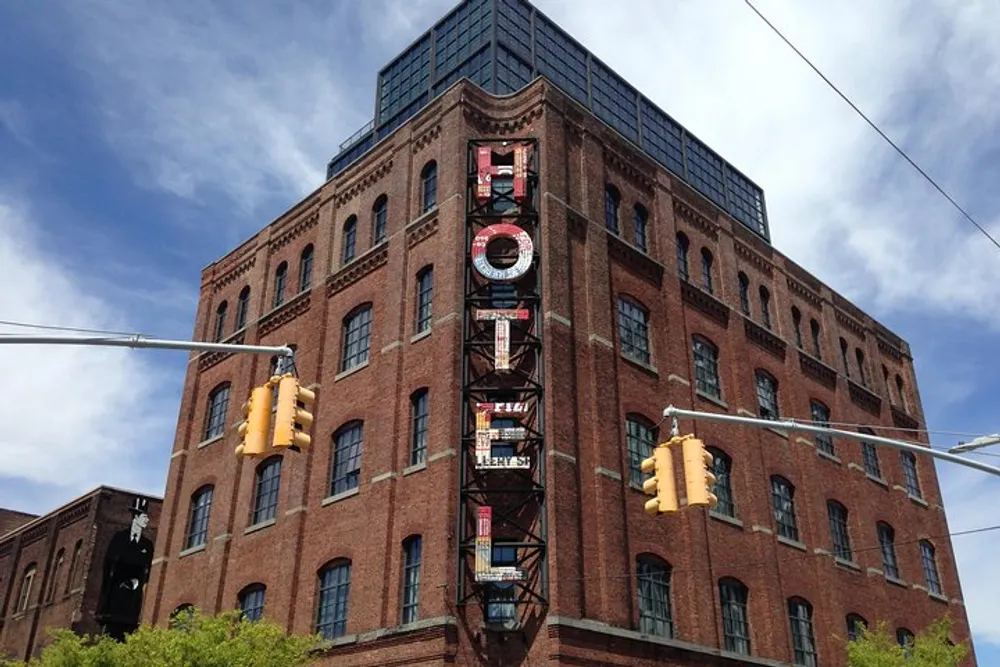 The image shows a tall brick building with a unique triangular shape featuring a fire escape and large letter signage on its side set against a blue sky with scattered clouds interspersed with traffic lights from an urban street perspective