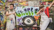 Two people are joyfully displaying their graffiti artwork featuring a large eye, with a dynamic and colorful backdrop full of street art.
