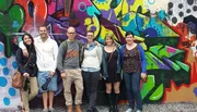 A group of smiling people is standing in front of a colorful graffiti-covered wall.