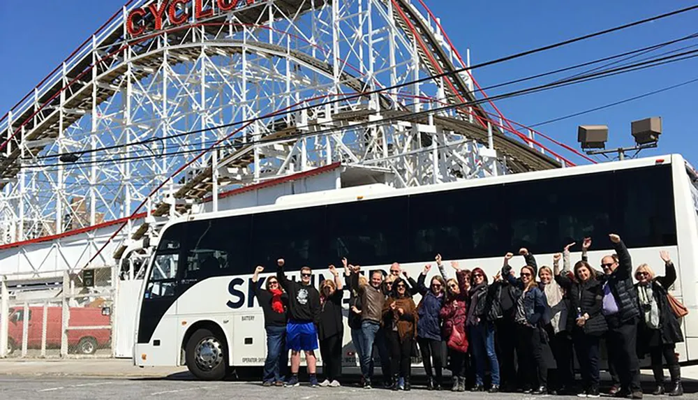A group of cheerful people are posing with raised arms in front of a parked bus with the historic wooden Cyclone roller coaster in the background