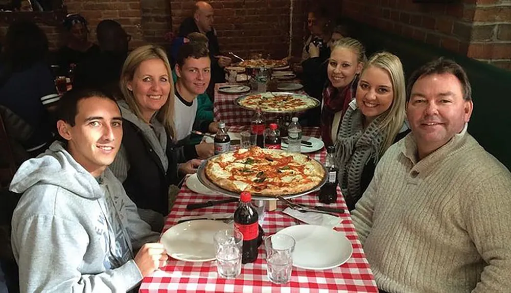 A group of six people is smiling at the camera while seated at a restaurant table covered with a red and white checkered tablecloth getting ready to enjoy a large pizza