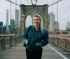 A smiling person in a denim jacket poses on the pedestrian walkway of the Brooklyn Bridge with the New York City skyline in the background