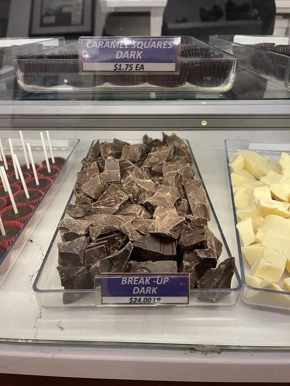 A tray filled with chunky pieces of dark chocolate labeled as Break-Up Dark for 2400 per pound is displayed in a candy store beside other confections