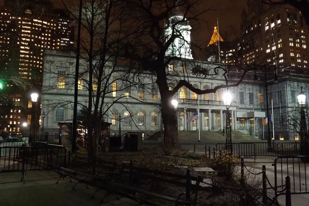 An evening view of a historic building framed by bare trees and surrounded by illuminated high-rise buildings