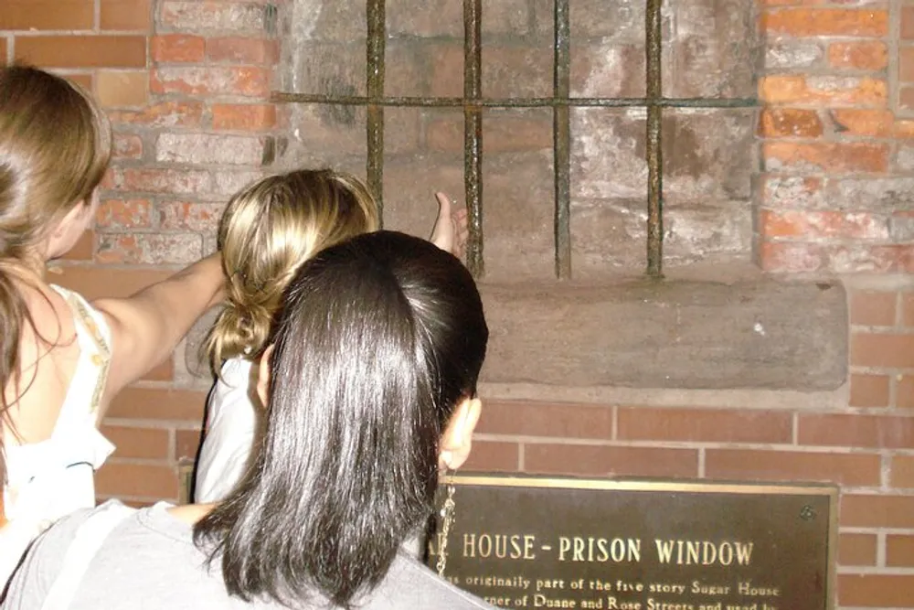 Two individuals are observing an old prison window behind bars on a brick wall with an informational plaque below it