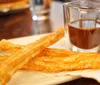 A plate of fresh churros is accompanied by a glass of chocolate sauce for dipping