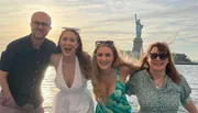 Four people are smiling and posing for a photo with the Statue of Liberty in the background, likely taken from a boat or waterfront in New York.