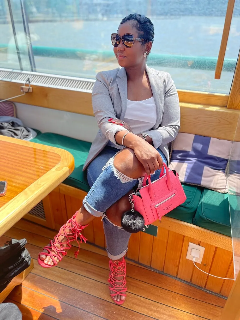 A person is sitting on a boat with stylish sunglasses a gray blazer ripped jeans bright pink strappy heels and a matching pink handbag while looking thoughtfully to the side