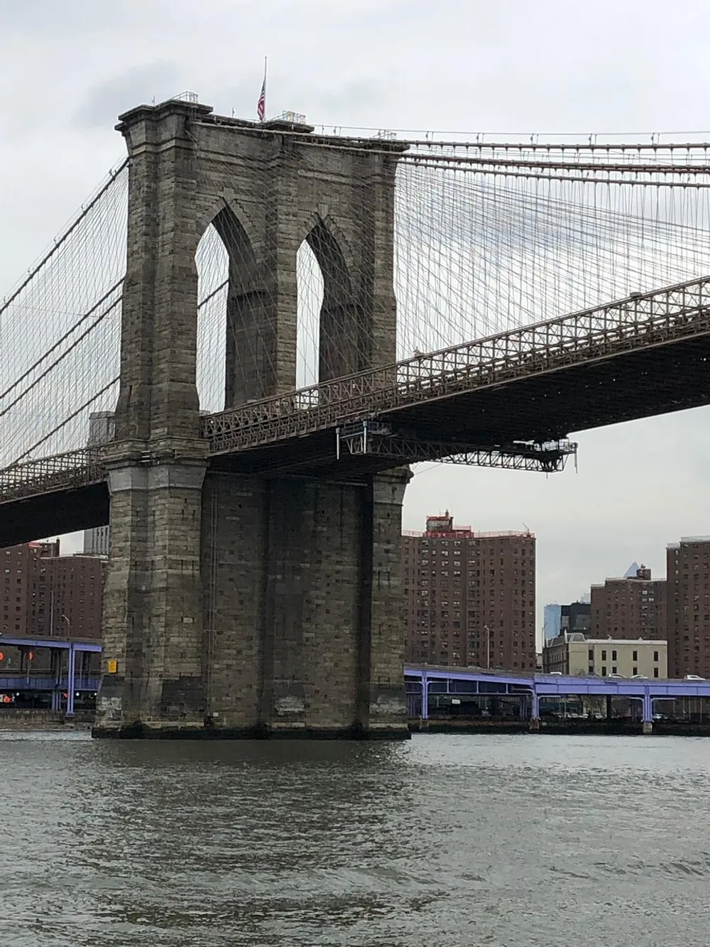 The image shows a close-up view of the Brooklyn Bridge tower and suspension cables over the East River with buildings visible in the background on a cloudy day