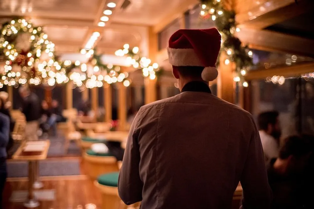 A person wearing a Santa hat is facing away from the camera overlooking a festive and warmly-lit interior adorned with holiday decorations