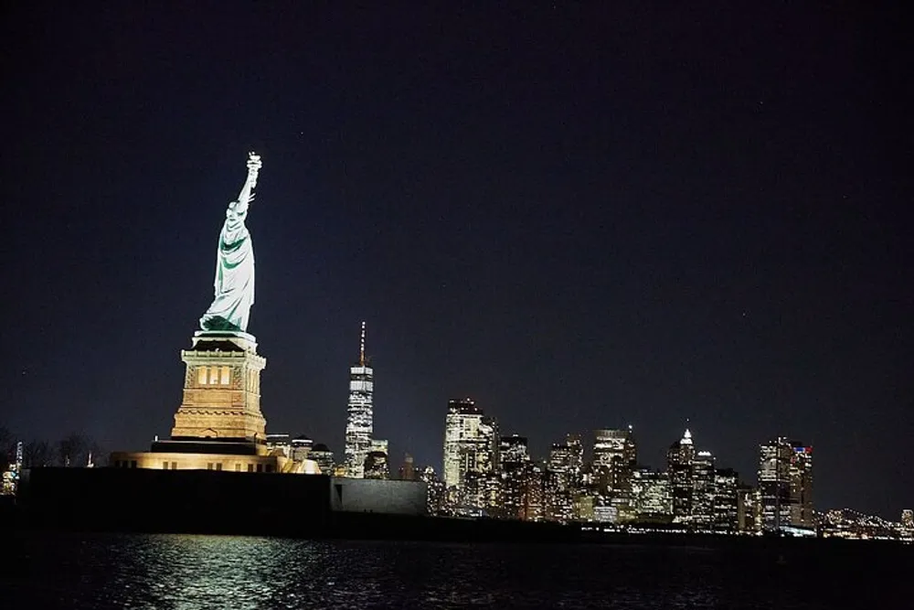 The Statue of Liberty is illuminated at night with the New York City skyline in the background