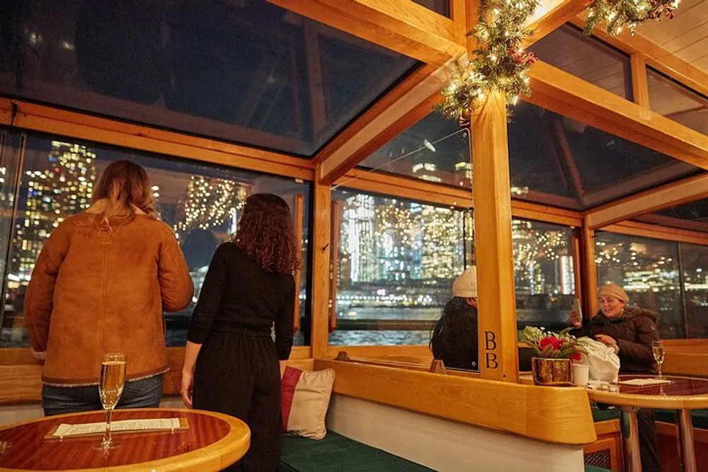 People enjoy the evening onboard a festively decorated boat with large windows offering a view of a citys sparkling night skyline