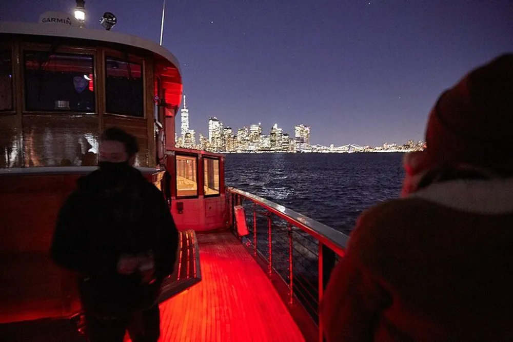 Passengers are standing on the deck of a boat at night enjoying the view of a brightly lit city skyline across the water