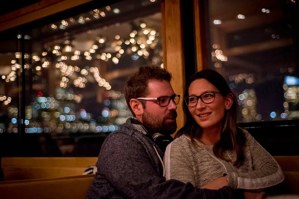 A man and a woman are sitting closely together smiling in a warmly lit setting with a backdrop of twinkling lights