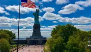The image shows the Statue of Liberty under a blue sky with scattered clouds, with an American flag in the foreground and visitors walking around the statue's pedestal.