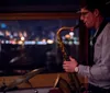 A saxophonist performs indoors at night with a backdrop of city lights through a window