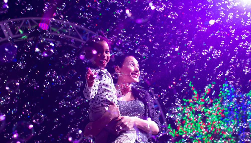 A joyful child is sitting on an adults shoulders both smiling and surrounded by a swirl of colorful bubbles against a backdrop of vibrant lights