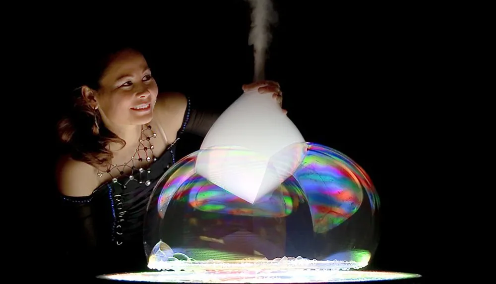 A person is smiling as they create large iridescent soap bubbles with visible smoke rising from a device in a dark setting