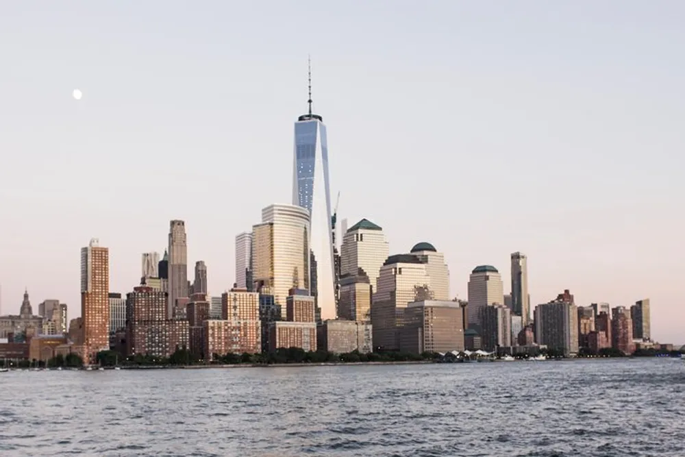 The image displays a scenic view of the Lower Manhattan skyline including the One World Trade Center seen from across the water during twilight with a clear sky and the moon faintly visible above