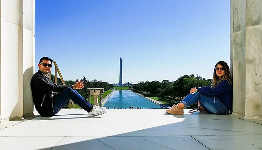 A man and a woman are sitting on the steps of the Lincoln Memorial with the Reflecting Pool and the Washington Monument in the background on a clear day