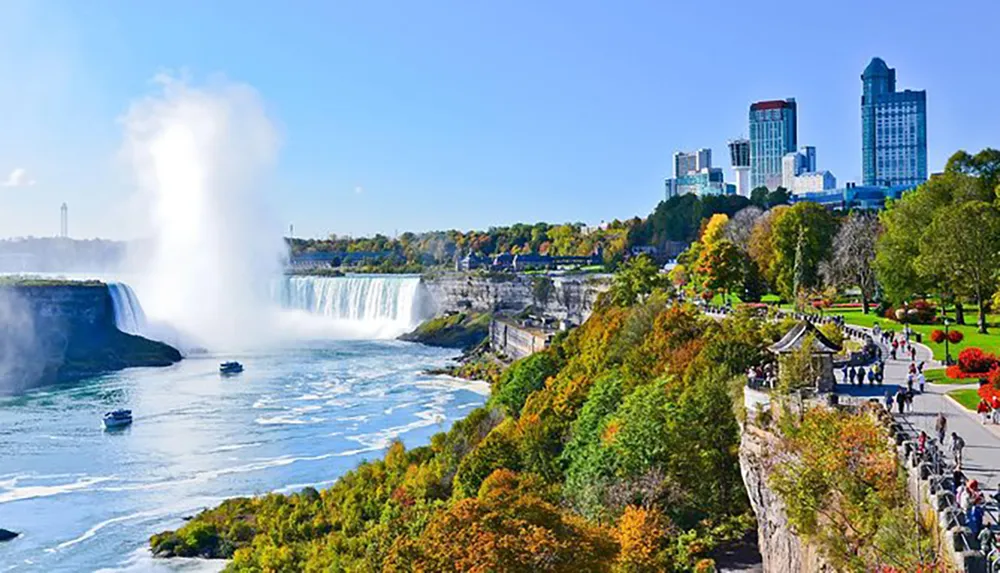 The image depicts the breathtaking Niagara Falls with mist rising from the cascading water tourist boats navigating the river and a bustling walkway lined with autumn-colored trees against a backdrop of a city skyline under a clear blue sky