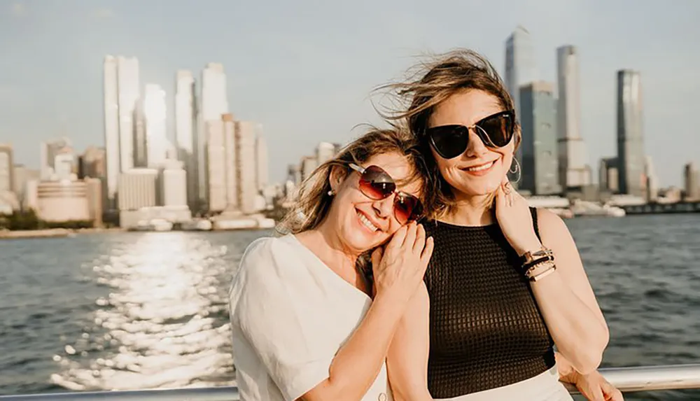 Two smiling women wearing sunglasses are posing for a photo with a city skyline in the background presumably taken from a boat on the water