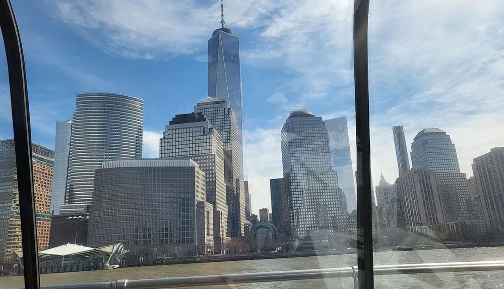 The photo captures a view of a dense cluster of skyscrapers including a very tall building taken from a position near or on water potentially through a glass barrier given the reflections and framing