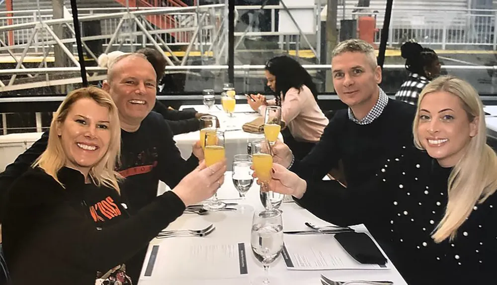 Four people are toasting with glasses of orange juice at a restaurant table smiling at the camera