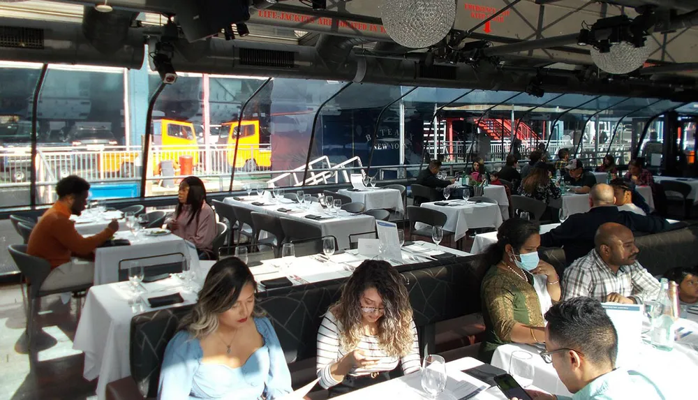 The image shows a group of people dining in an elegant indoor setting with large windows offering a view of a city bus outside and some patrons appearing engaged in conversation or checking their phones