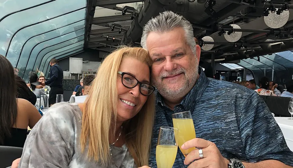 A smiling couple is posing for a photo while holding glasses of a yellow beverage in a dining area with a modern glass enclosure