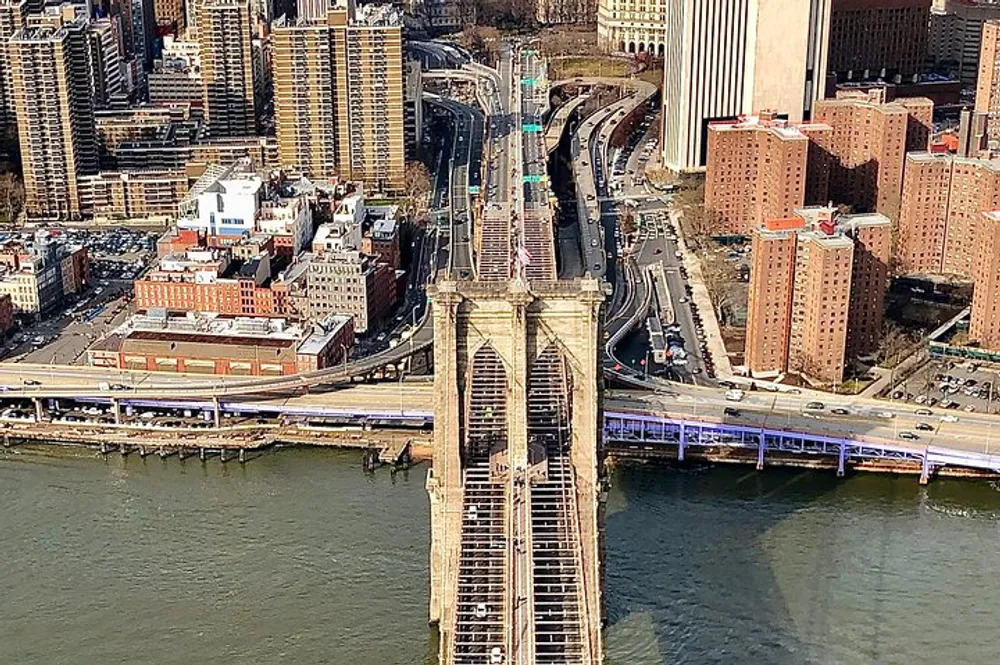 The image shows an aerial view of the Brooklyn Bridge spanning over the East River with cars on the roadway and surrounded by the urban landscape of New York City