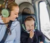 A woman and a young child are smiling and wearing headphones while sitting inside a helicopter with a cityscape visible through the window