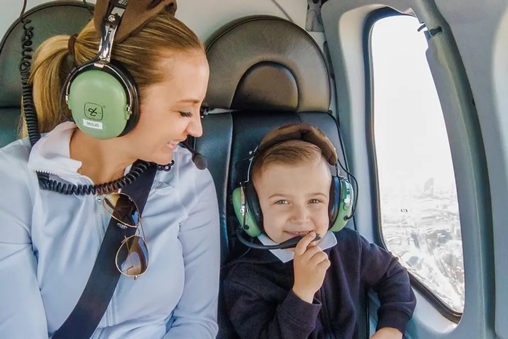 A woman and a young child are smiling and wearing headphones while sitting inside a helicopter with a cityscape visible through the window