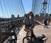 Two cyclists are standing near their bikes along a riverside with a bridge and the city skyline in the background under a clear blue sky