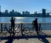 Two cyclists are standing near their bikes along a riverside with a bridge and the city skyline in the background under a clear blue sky