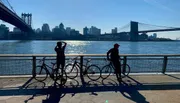 Two cyclists are standing near their bikes along a riverside, with a bridge and the city skyline in the background, under a clear blue sky.