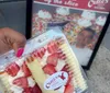 A person is holding a slice of strawberry-topped cake in a clear container in front of a Carlos Bakery sign in Hoboken New Jersey
