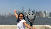 A person poses with an outstretched arm, appearing to touch the top of a skyscraper in the distance, with a view of a city skyline and a coin-operated binocular in the foreground.