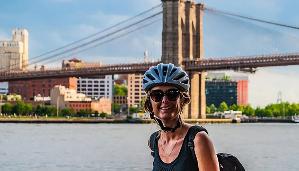 A woman wearing a bicycle helmet smiles in front of the Brooklyn Bridge and the East River