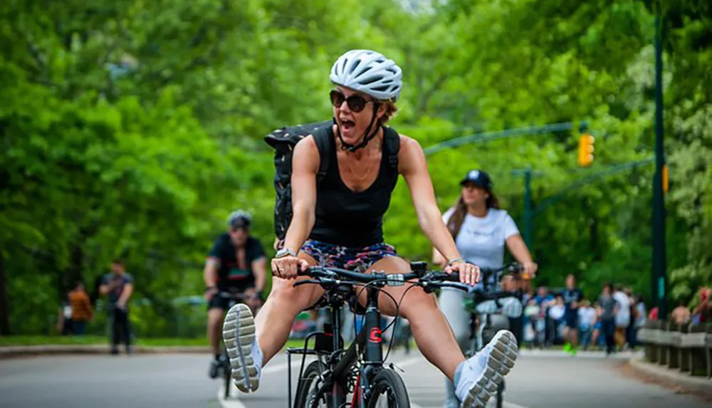 A person is energetically riding a bicycle with their feet off the pedals and mouth open wide as if shouting while others cycle and walk in the background