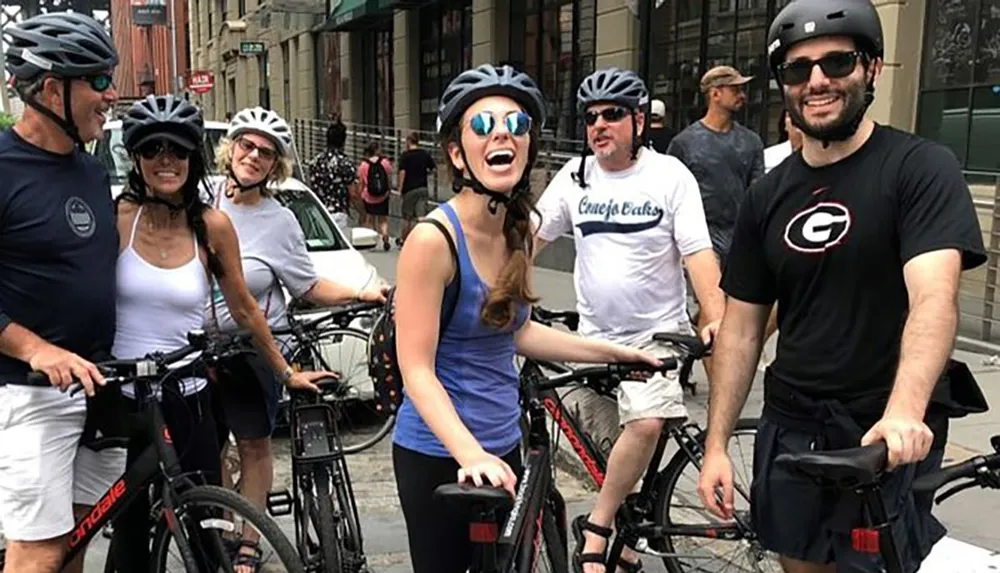 A group of smiling people wearing helmets are standing with their bicycles on an urban street