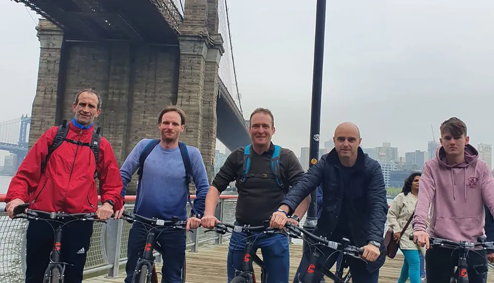 Five people are standing with their bikes on a walkway with a suspension bridge visible in the background